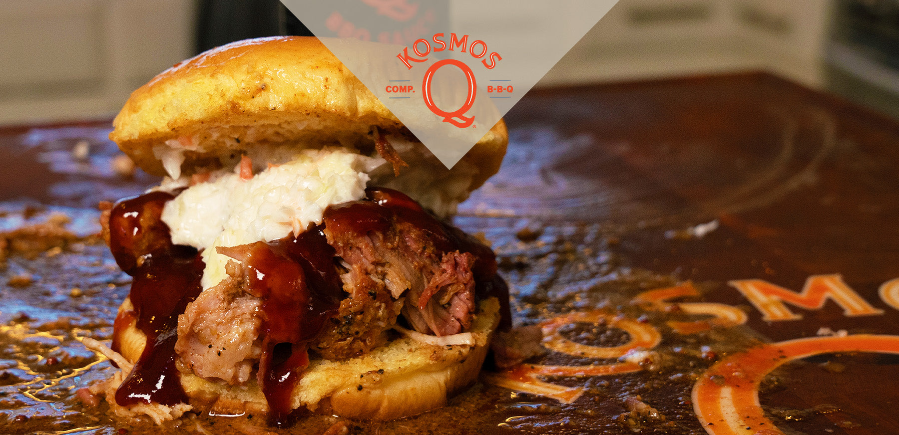 Close up of a pork slider, heaping with juicy pulled pork, topped with BBQ sauce and coleslaw. It is placed on a cutting board with part of the Kosmos Q logo visible. The logo is also overlayed in the top center of the image.